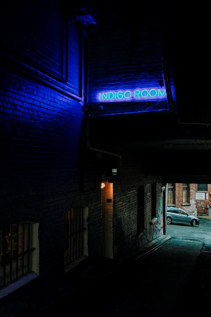 A neon blue light saying Indigo Room shines on a street art-adorned alley.