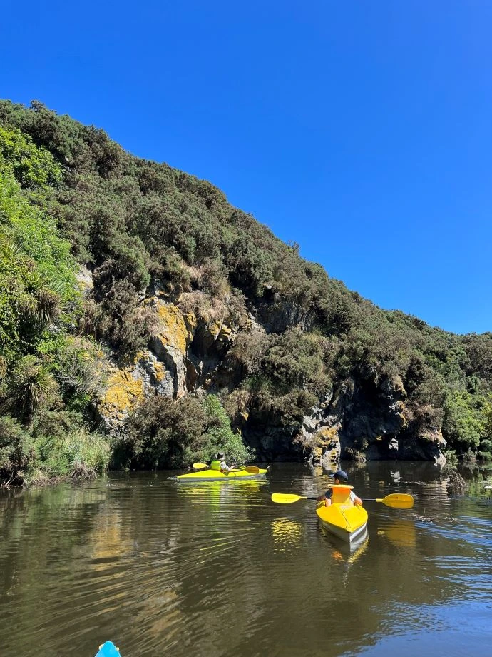 Two kayakers on the creek paddle towards a rocky mound.