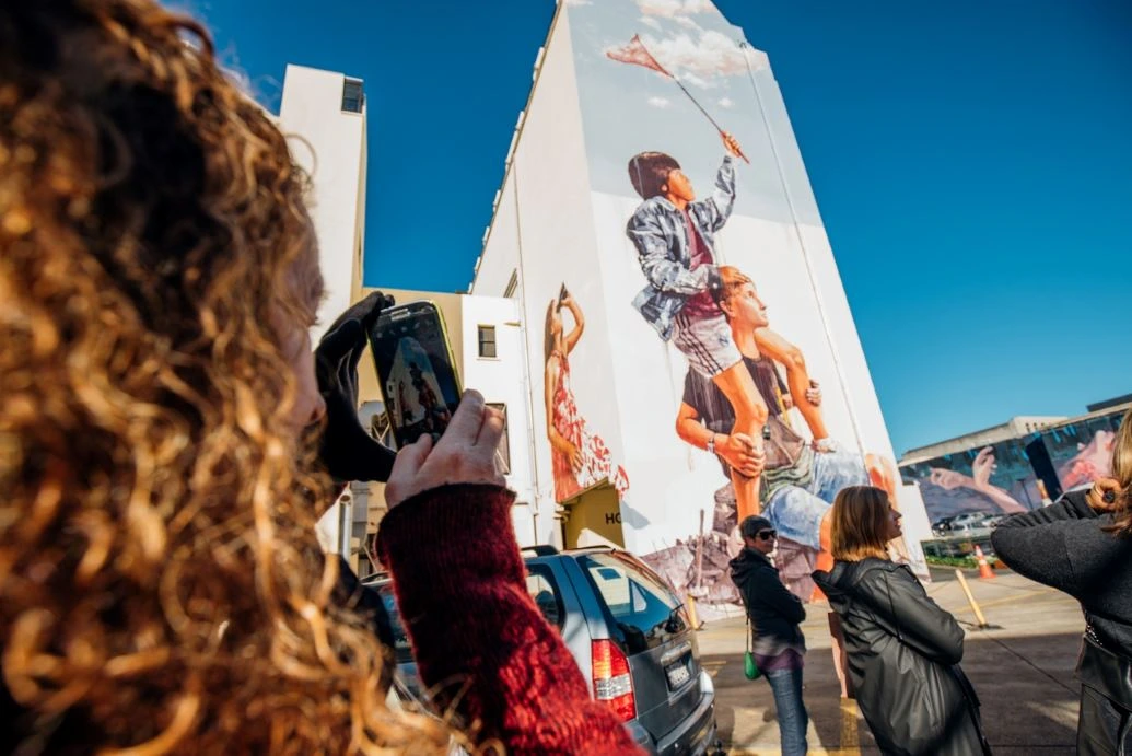 People take photos of a large mural depicting children catching clouds with a butterfly net.