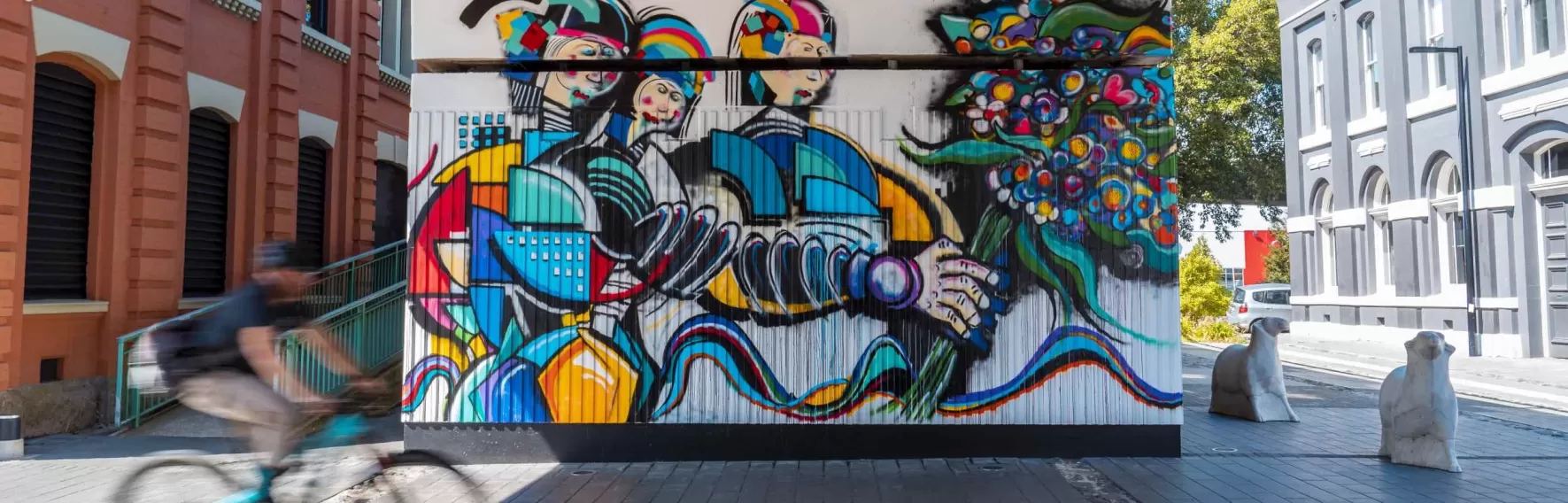 A cyclist speeds past some colourful street art on a wall beneath an overpass.