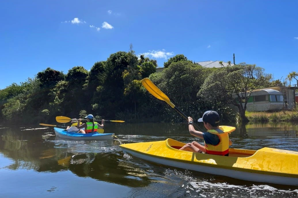 Children paddle blue and yellow kayaks down a creek.