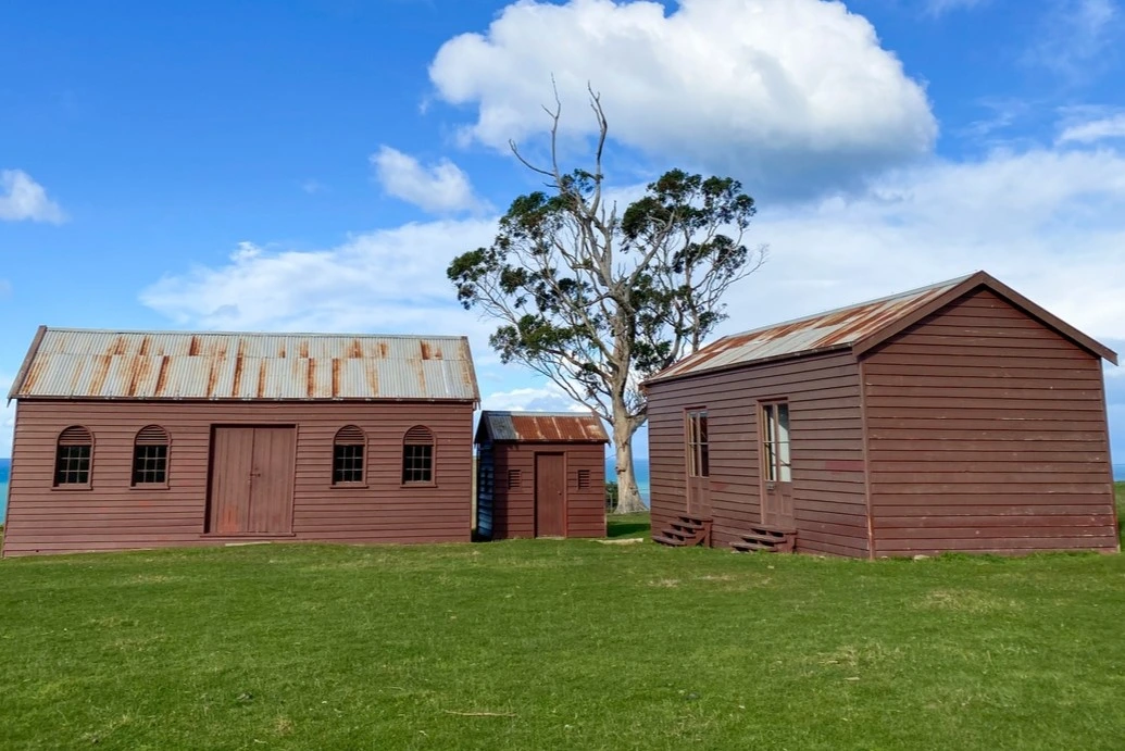 Three red-brown heritage structures, the original settler farm buildings stand on a green lawn.